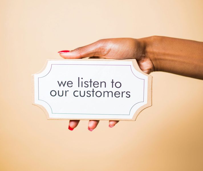 A hand holding a sign saying 'we listen to our customers' on a neutral background.